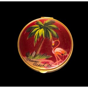 May include: A circular vintage compact with a red background and a gold rim. It features a hand-painted scene of a palm tree, a flamingo, and the words "Miami Beach Fla."