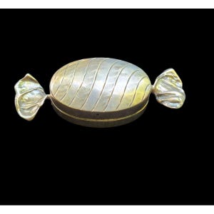 May include: A metallic, candy-shaped box with a polished, oval-shaped center featuring diagonal etched lines. The ends of the object are sculpted to resemble the twisted wrappers of a piece of hard candy, finished in a smooth, reflective silver tone.