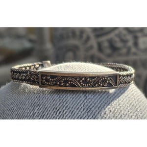 Può includere: Un bracciale d'argento con un elemento centrale rettangolare ornato e un design a catena intrecciata. L'elemento centrale presenta un motivo dettagliato di linee a spirale e piccoli punti. Il bracciale è esposto su una superficie di denim.