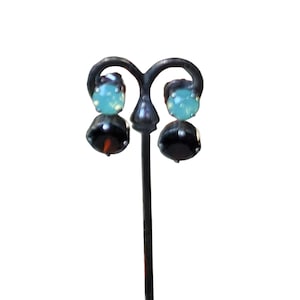 May include: A pair of earrings with a two-tone design. Each earring has a light blue, round top and a black, faceted bottom. The earrings are displayed on a black metal stand against a white background.