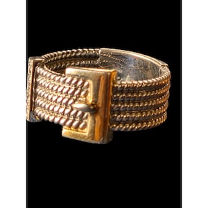 May include: A gold-toned ring with a textured design. The ring features a rectangular buckle-like element and multiple rows of twisted rope-like detailing. The ring is set against a black background.