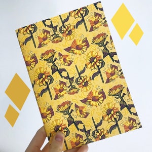 May include: A notebook with a yellow cover featuring a repeating pattern of sunflowers in shades of yellow, orange, and brown. The sunflowers have detailed petals and dark green stems. The background is a lighter yellow with diamond shapes.