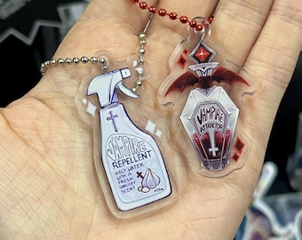 Peut inclure: Deux breloques en acrylique avec des attaches à chaîne à billes. L'une est un flacon pulvérisateur avec l'inscription "Vampire Repellent" et une illustration de croix et d'ail. L'autre est une bouteille en forme de cercueil avec l'inscription "Vampire Attractor" et des ailes de chauve-souris.