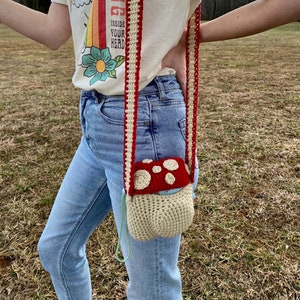 May include: A crocheted mushroom-shaped bag with a red top and white spots. The bag is attached to a red and white striped crocheted strap. The bag is being worn by a person wearing blue jeans.