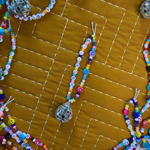 May include: Colorful beaded necklaces with smiley faces, stars, and letter beads spelling out words like "SUGAR" and "ARIES". Each necklace features a disco ball pendant. The beads are in various colors, including pink, blue, yellow, and green.