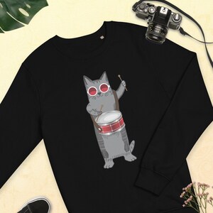 May include: Black sweatshirt with a gray cat wearing red sunglasses and playing a drum. The cat is holding a drumstick in its right paw and has a red drum in front of it.