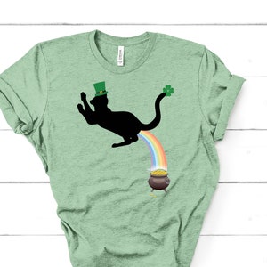 May include: A green t-shirt with a black cat wearing a green hat and a shamrock on its tail. The cat is pooping a rainbow that leads to a pot of gold.