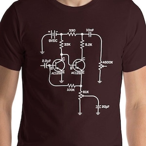 以下が含まれることがあります： 電子回路図が白で描かれた、マルーンのTシャツ。回路図には抵抗器、コンデンサ、トランジスタが含まれています。回路図のテキストには、330、10nF、SVDC、33K、8.2K、2.2µF、4500K、AC128E、200K、BIK、20µFが含まれています。