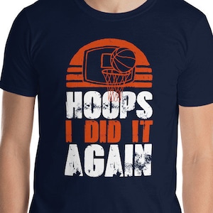 Könnte beinhalten: Marineblaues T-Shirt mit Basketballkorb-Grafik und dem Text "HOOPS I DID IT AGAIN" in Weiß und Orange. Das Design zeigt einen Basketball und einen sonnenuntergangsähnlichen Hintergrund.