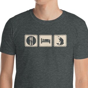 Puede incluir: Una camiseta gris oscuro jaspeada con un gráfico de tres iconos: un tenedor y un cuchillo, una cama y un pez. El texto "Comer, Dormir, Pescar" está impreso debajo de los iconos.