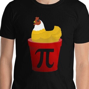 May include: Black t-shirt featuring a cartoon chicken inside a red bucket with the pi symbol. The chicken has yellow feathers and a red comb. The shirt is made of a soft material.