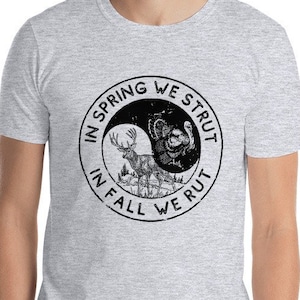 Könnte beinhalten: Ein heidefarbenes T-Shirt mit einem schwarzen Yin-Yang-Symbol. Die linke Seite des Symbols lautet "In Spring We Strut" und die rechte Seite lautet "In Fall We Rut".
