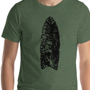 Puede incluir: Camiseta verde con un diseño abstracto grande en negro. El diseño se asemeja a una punta de flecha o un artefacto antiguo. La camiseta está hecha de un material suave y cómodo. La camiseta es de manga corta.