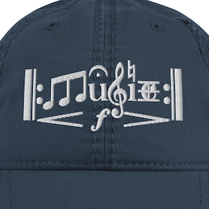 Può includere: Un cappellino da baseball blu navy con effetto invecchiato, con note musicali ricamate in bianco e la parola "music". Il cappellino ha una visiera curva e un aspetto vintage.