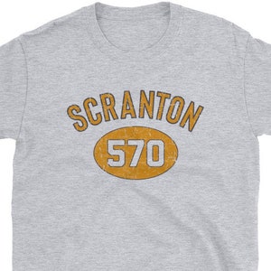 May include: Gray short-sleeved t-shirt with a vintage-style design featuring the text "SCRANTON 570" in a yellow oval.
