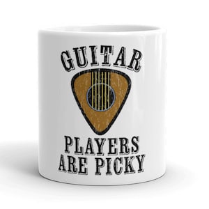 Può includere: Tazza da caffè in ceramica bianca con testo nero che recita "Guitar Players Are Picky". Un plettro per chitarra marrone con contorno nero è sopra il testo.