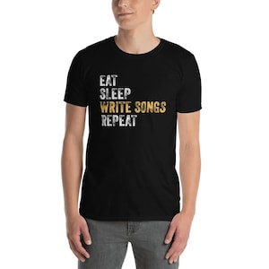 Puede incluir: Camiseta negra con texto blanco que dice "Eat Sleep Write Songs Repeat" en una fuente desgastada. La palabra "Write Songs" est&aacute; en amarillo.