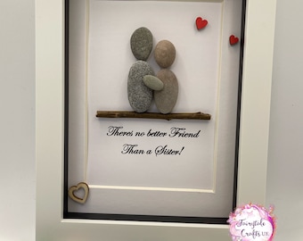 May include: A framed pebble art picture with two figures holding each other. The figures are made of gray and brown pebbles. The background is white with the text "There's no better Friend Than a Sister!" and three red hearts. The frame is white with a black mat.