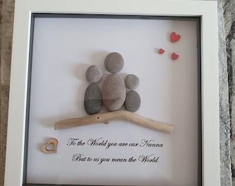 May include: A framed pebble art piece with a family of three represented by gray stones sitting on a driftwood branch. The frame has a white mat and a white frame. The background is white with the text "To the World you are our Nanna But to us you mean the World" and three red hearts.