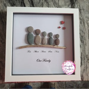 May include: A framed pebble art family portrait with five figures standing on a driftwood branch. The names Tom, Alison, Anna, Sam, and Nora are written below the branch. The words "Our Family" are written below the names. There are three red hearts in the top right corner of the frame.