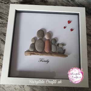 May include: A framed pebble art picture of a family with a dog. The family is made of gray, brown, and red stones. The dog is made of gray stones. The picture is on a white background with the word "Family" written in black below the family. The picture is framed in white with a gray mat.