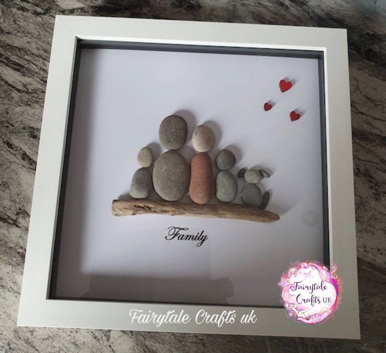 May include: A framed pebble art picture of a family with a dog. The family is made of gray, brown, and red stones. The dog is made of gray stones. The picture is on a white background with the word "Family" written in black below the family. The picture is framed in white with a gray mat.