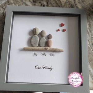 May include: A framed pebble art piece depicting a family of three with the names Gaz, Abby, and Elsie written below. The artwork is on a white background with red hearts in the top right corner and the words "Our Family" written below the names. The frame is gray.