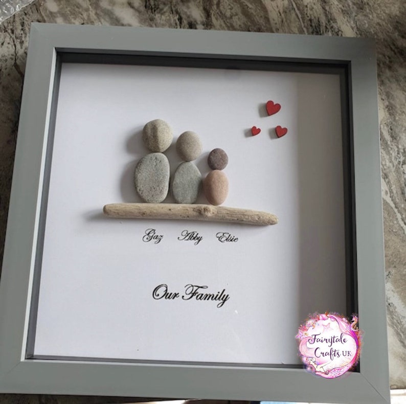 May include: A framed pebble art piece depicting a family of three with the names Gaz, Abby, and Elsie written below. The artwork is on a white background with red hearts in the top right corner and the words "Our Family" written below the names. The frame is gray.