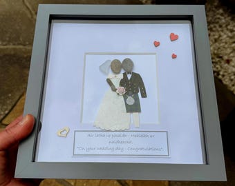 May include: Framed wedding art featuring a bride and groom made of pebbles. The bride wears a white dress and veil, holding a pink flower. The groom wears a black suit and kilt. The frame is gray, with small heart accents and text.