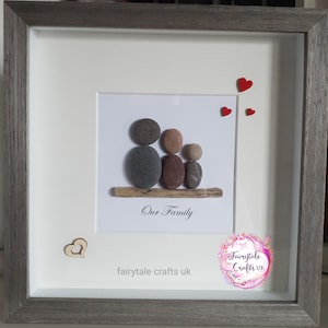 May include: A framed pebble art piece depicting a family of three. The pebbles are arranged to look like a family sitting on a piece of driftwood. The frame is gray with a white mat. The text "Our Family" is written below the pebble art. The text "fairytale crafts uk" is written below the text "Our Family". There are three red hearts in the top right corner of the image.
