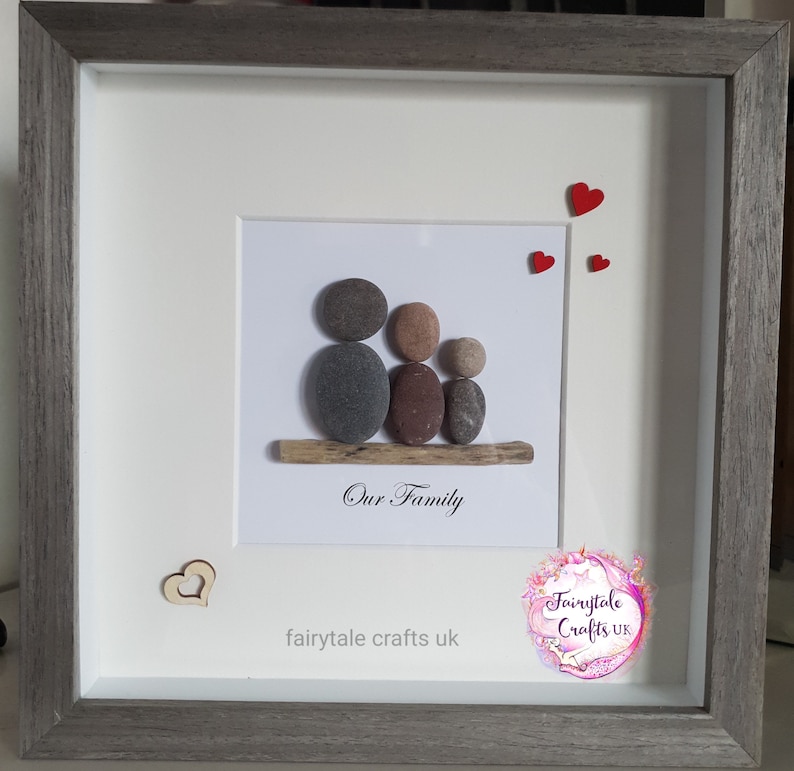 May include: A framed pebble art piece depicting a family of three. The pebbles are arranged to look like a family sitting on a piece of driftwood. The frame is gray with a white mat. The text "Our Family" is written below the pebble art. The text "fairytale crafts uk" is written below the text "Our Family". There are three red hearts in the top right corner of the image.