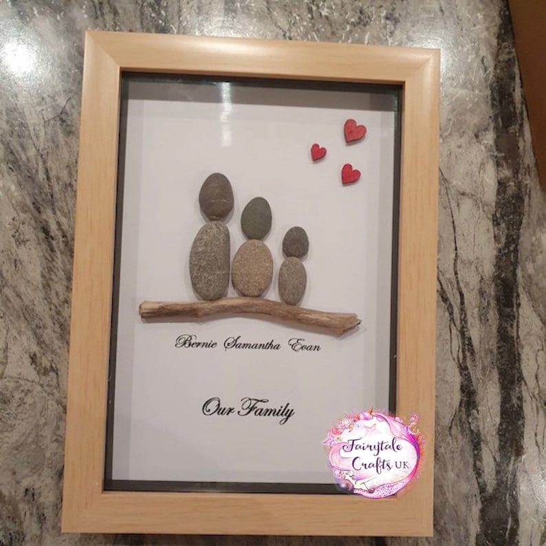May include: A framed pebble art piece depicting a family of three with the names Bernie, Samantha, and Evan written below. The artwork includes a small twig and three red hearts. The bottom of the frame reads "Our Family" and the bottom right corner has a logo that reads "Fairytale Crafts UK".