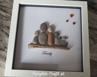 May include: A framed pebble art piece depicting a family of five with a dog. The family is sitting on a piece of driftwood with the word "Family" written below them. There are three red hearts in the top right corner of the frame.