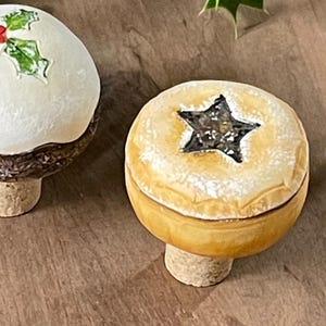 May include: Two festive Christmas ornaments are shown. One is a white, round ornament decorated with green holly and red berries. The other is a yellow pie-shaped ornament with a star design. Both ornaments have a cork-like base.