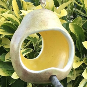 May include: A ceramic bird feeder shaped like a pear with a yellow glazed interior and a small twig attached to the bottom. The feeder is hanging from a string.