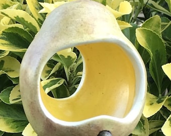 May include: A ceramic bird feeder shaped like a pear with a yellow glazed interior and a small twig attached to the bottom. The feeder is hanging from a string.