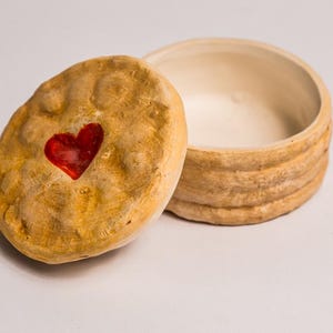 Może przedstawiać: Małe ceramiczne pudełko na biżuterię z pokrywką. Pokrywka jest jasnobrązowa z czerwonym sercem. Okrągłe pudełko ma żebrowaną fakturę i kremowe wnętrze. Wymiary w centymetrach.