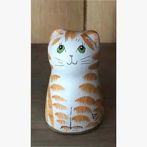 May include: A hand-painted ceramic cat figurine. The cat is white with orange tabby markings and green eyes. The figurine is standing upright with a simple, stylized design. The cat has a smiling face and is a decorative item.