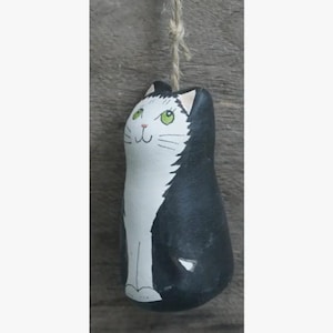 May include: A hand-painted, black and white cat ornament with green eyes. The cat is hanging from a natural twine string. The ornament has a whimsical, folk art style and is suitable for cat lovers.