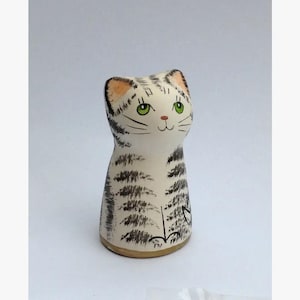 May include: A cream-coloured ceramic cat figurine with black stripes. The cat has green eyes, a pink nose, and a smiling mouth. The ears are a light orange colour. The figurine is sitting upright.