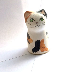 May include: A ceramic figurine of a calico cat with green eyes. The cat is sitting with its legs tucked under its body. The figurine is painted in shades of white, black, and orange.