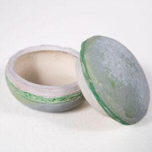 May include: A small, round ceramic trinket box with a lid. The box is painted in shades of grey and green with a white rim. The lid has a similar design and is slightly domed.