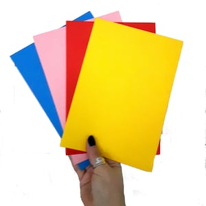 Puede incluir: Cuatro hojas de papel de colores, azul, rosa, rojo y amarillo, est&aacute;n apiladas y sostenidas en una mano.