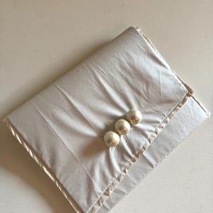 Può includere: Un tessuto beige chiaro piegato con un aspetto satinato. Tre grandi bottoni rotondi a forma di perla sono allineati nell'angolo in alto a destra. Il tessuto ha una trama sottile e un bordo piegato.