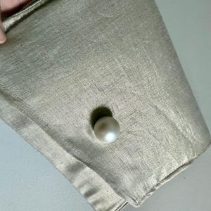 Può includere: Primo piano di un tessuto beige chiaro con un bottone a forma di perla. Il tessuto ha una lucentezza sottile e un aspetto strutturato. Il bottone è rotondo e ha una finitura lucida, in contrasto con il tessuto opaco.