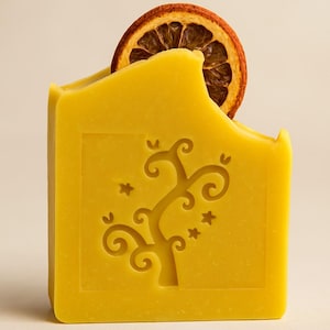 Könnte beinhalten: Ein gelbes, rechteckiges Seifenstück mit einem geschnitzten Baumdesign und einer getrockneten Orangenscheibe. Die Seife hat eine gewölbte Oberseite und ein dekoratives Baum- und Sternmuster. Die Orangenscheibe ist braun.