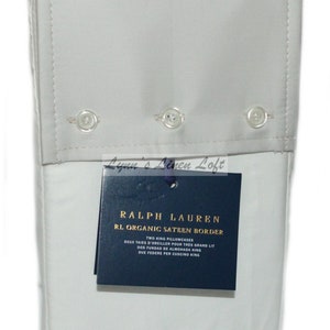 May include: A folded, light gray pillowcase set with a buttoned border. The tag reads "RALPH LAUREN RL ORGANIC SATEEN BORDER" and lists the product as two king pillowcases. The set is neatly folded and appears to be made of a soft fabric.
