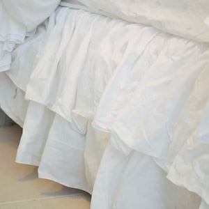 May include: White bed skirt with a ruffled edge. The skirt is made of a soft, flowing fabric and has a simple, elegant design.