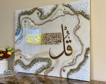 May include: A rectangular wall art piece with a white and gold abstract design. The artwork features gold Arabic calligraphy and is framed with a border of white and gold crystals. The piece is displayed on a dark brown surface.