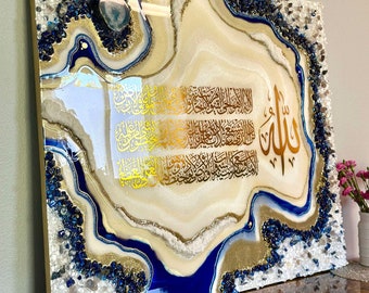 May include: A decorative wall art piece featuring Arabic calligraphy in gold against a cream and blue agate-style background. The artwork is framed with blue and clear crystals, and gold accents. The overall design is elegant and spiritual.